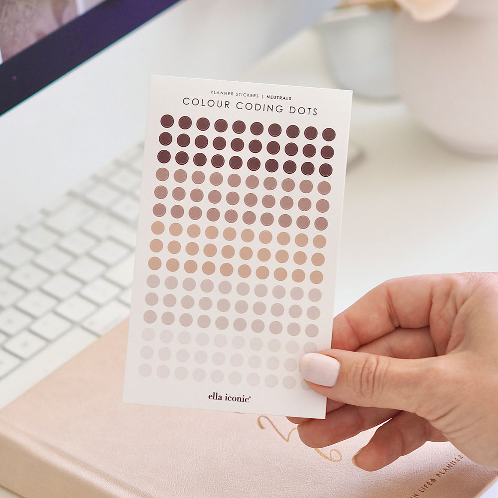 Hand holding a sheet of Colour Coding Dots by ella iconic on a desk with a keyboard and notebook.